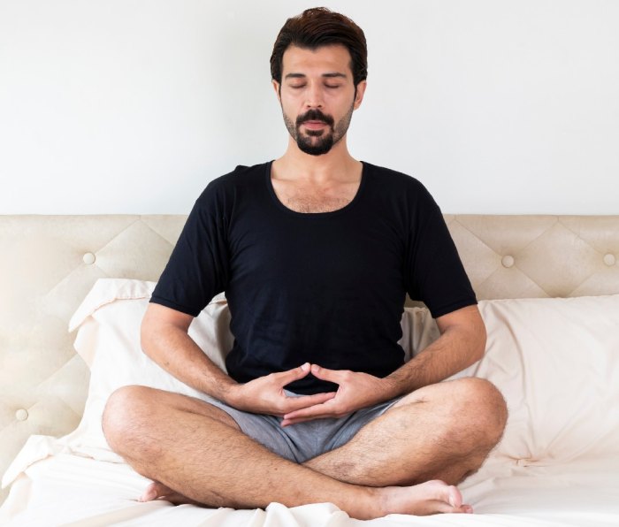 Человек, сидящий со скрещенными ногами на кровати, делает дыхательные упражнения